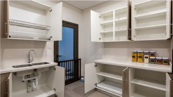 An empty kitchen in a villa prepared for pest control, with open cabinets, cleared countertops, an empty under-sink cupboard, sealed food containers, and a closed pet gate in the background.